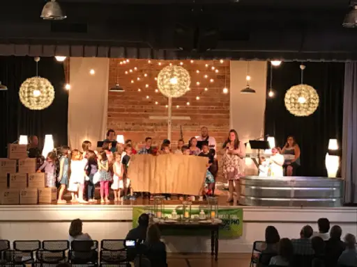 Children perform on stage as adults and an audience watch. The event features a display about plastic pipes from Titan Plumbing & Drains.